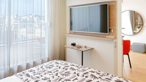 Habitación de hotel con una cama grande, un televisor montado en la pared, una mesita con una cámara, un espejo y vistas a la ciudad por la ventana.