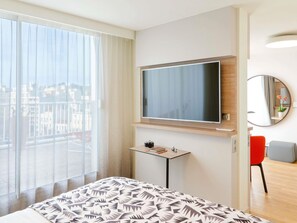 A hotel room with a large bed, a TV mounted on the wall, a small table with a camera, a mirror, and a view of the city through the window. - Mercure Marseille Centre Prado Vélodrome (Marseille)
