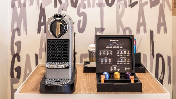 Nespresso-kahvinkeitin ja kahvikapselien pidike puisella pöydällä, taustalla seinä, jossa on suuria kirjaimia.