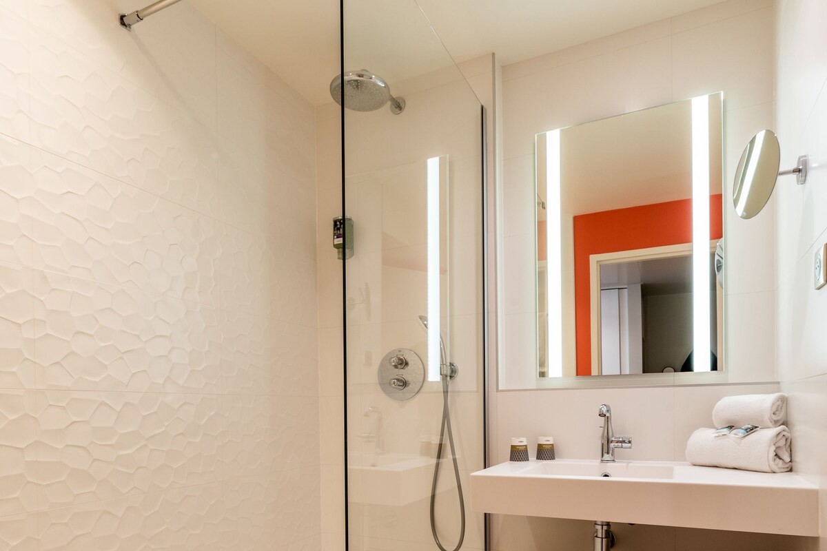 A modern bathroom with a glass-enclosed shower, a white sink with a wooden shelf, and a mirror with a red accent wall.