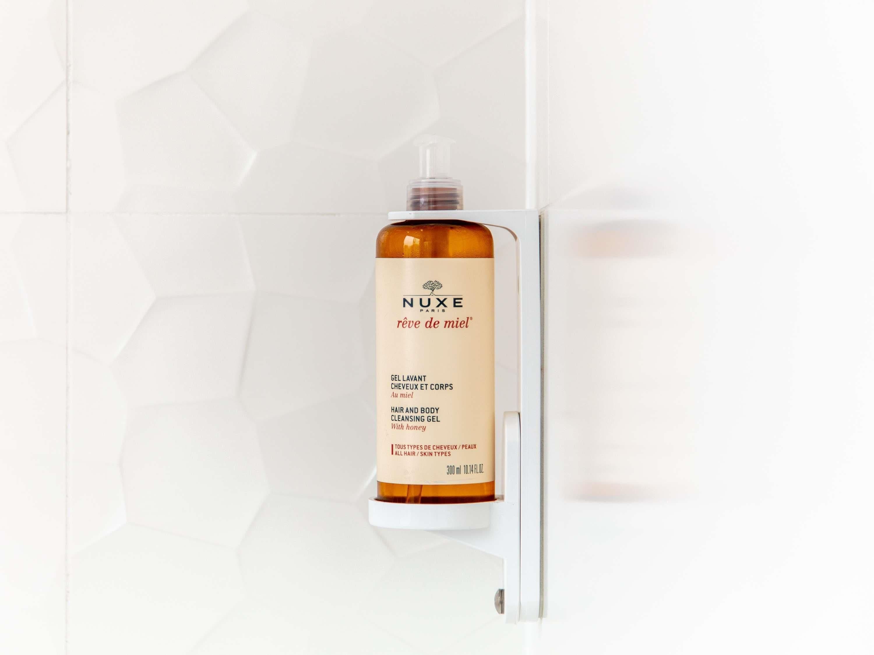 A bottle of Nuxe body wash on a white tiled wall.