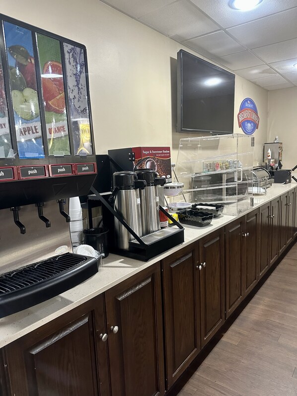 Estación de desayuno con máquina de café, dispensador de azúcar y edulcorantes, y dispensador de bebidas que ofrece opciones de manzana, naranja y agua.