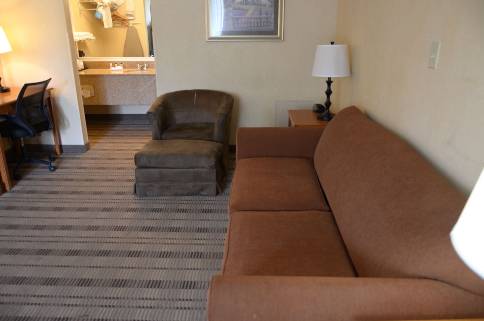 Ein Wohnzimmer mit braunem Sofa, einem braunen Sessel, einem kleinen Hocker, einem Schreibtisch mit Stuhl, einer Lampe und einem gerahmten Bild an der Wand.