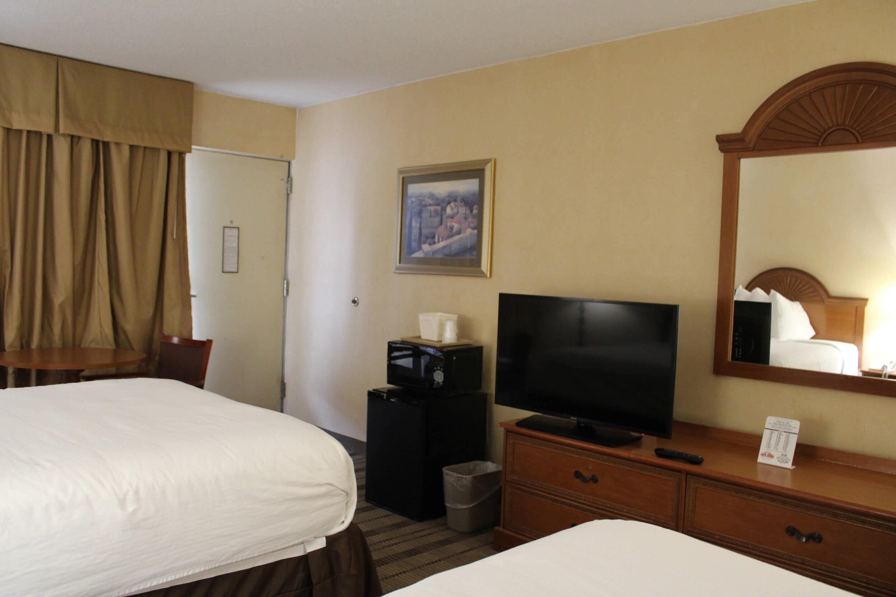 A hotel room with two beds, a television, a small refrigerator, a trash bin, and a mirror.
