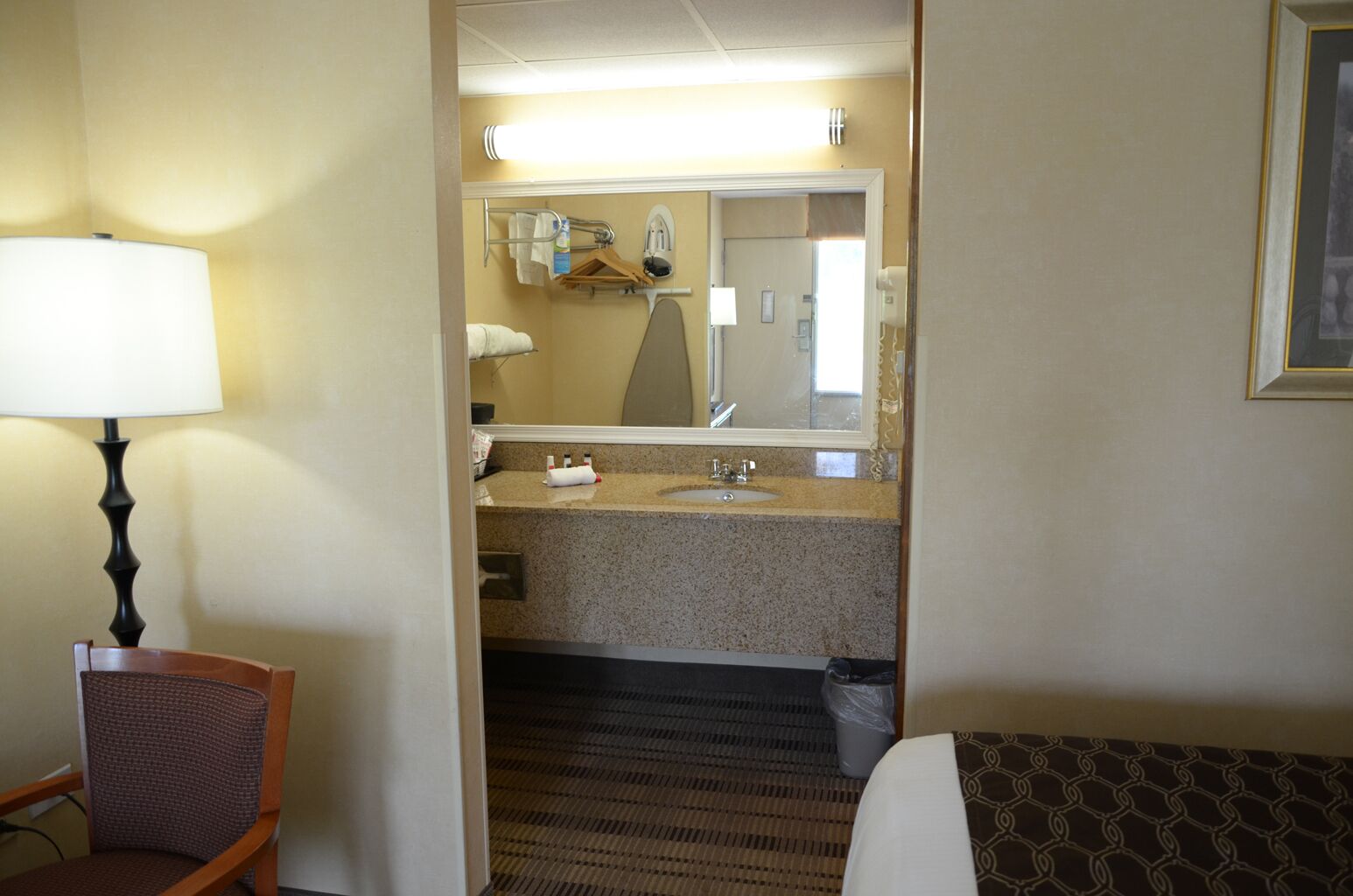 A hotel room with a bed, a chair, a desk, a lamp, a bathroom with a sink and mirror, and a trash bin.