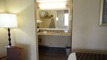 A hotel room with a bed, a chair, a desk, a lamp, a bathroom with a sink and mirror, and a trash bin.