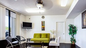 A living room with a green sofa, two black chairs, a coffee table, a flat-screen TV, a painting, a potted plant, and a door. - Motel 6 Covington, TN (Covington)