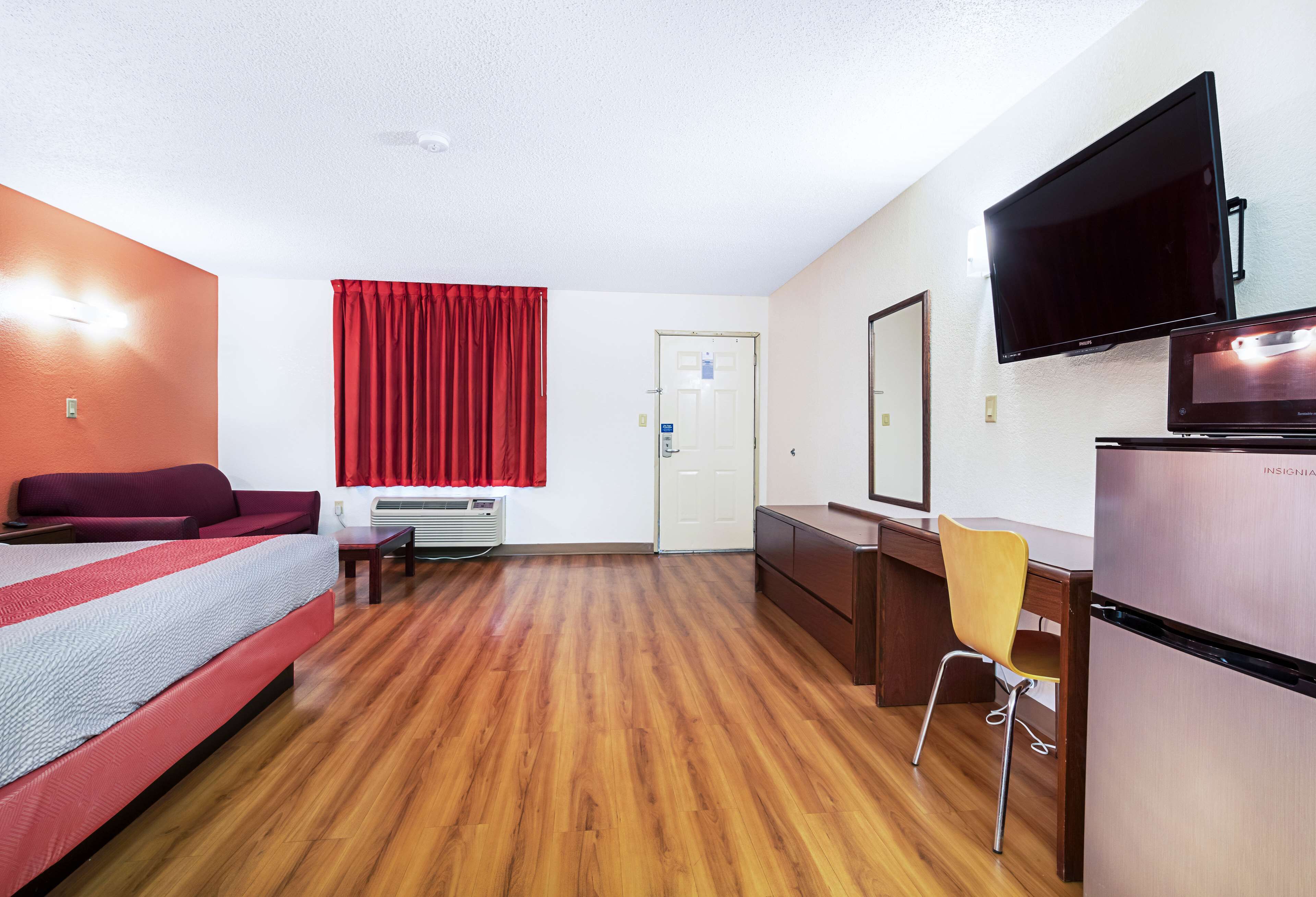 A hotel room with a bed, a desk with a chair, a flat-screen TV, a refrigerator, and a window with red curtains.