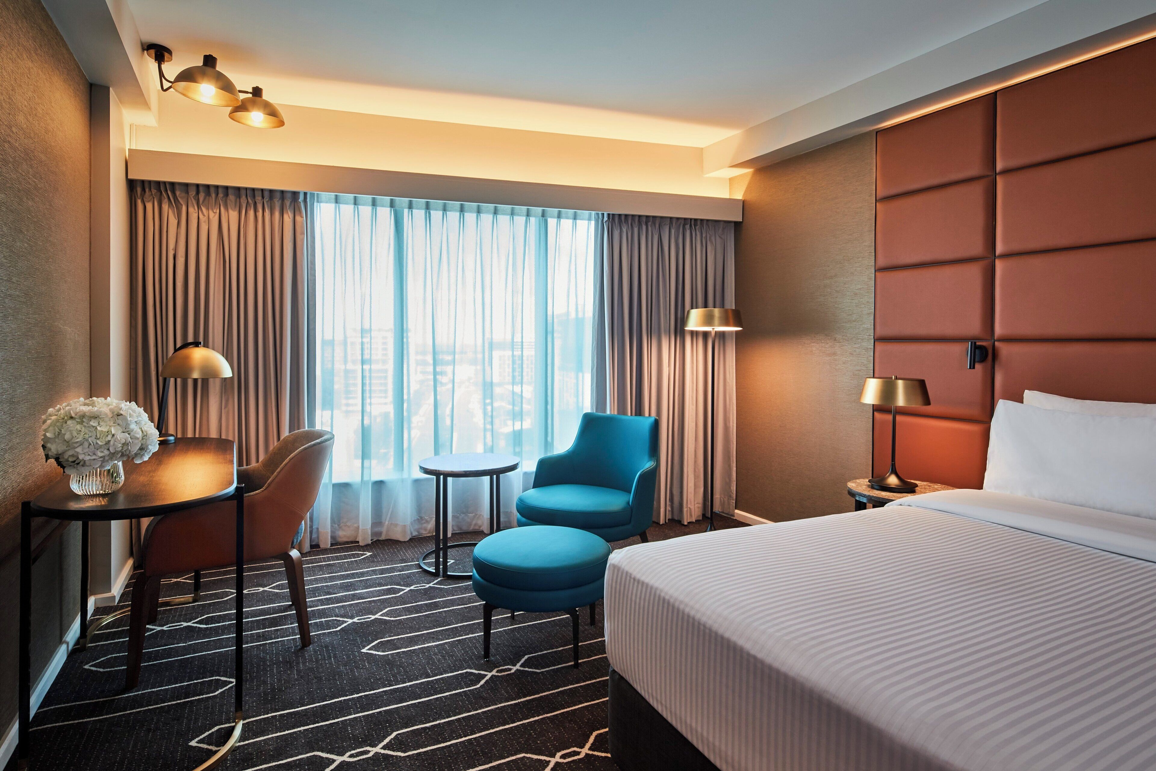 A hotel room with a bed, two chairs, a small table, a lamp, and a view of the city through large windows.