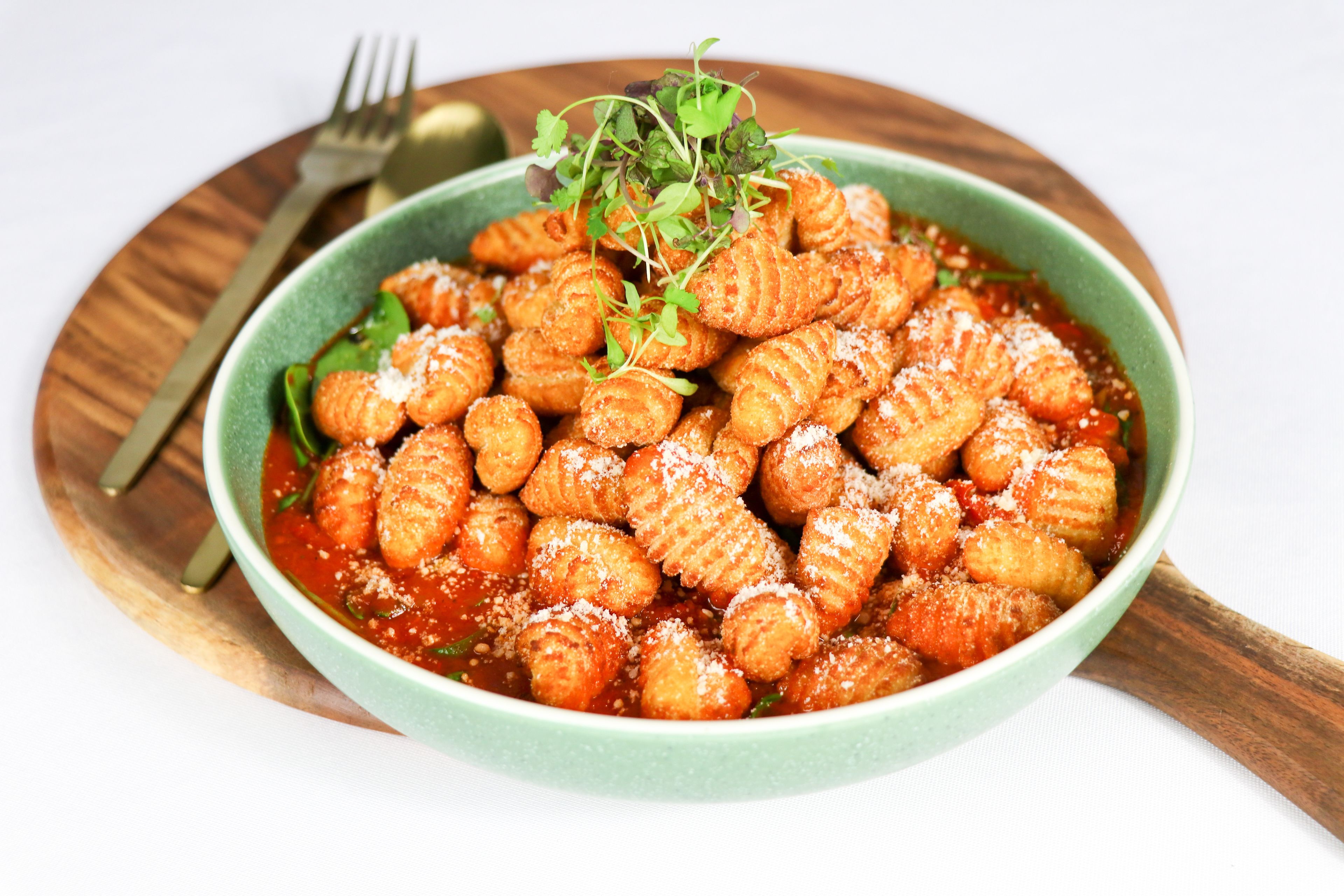 A bowl of ravioli with a garnish of greens and grated cheese, served on a wooden plate with a fork and spoon beside it.