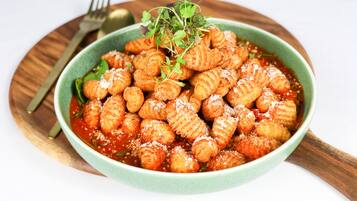 A bowl of ravioli with a garnish of greens and grated cheese, served on a wooden plate with a fork and spoon beside it.