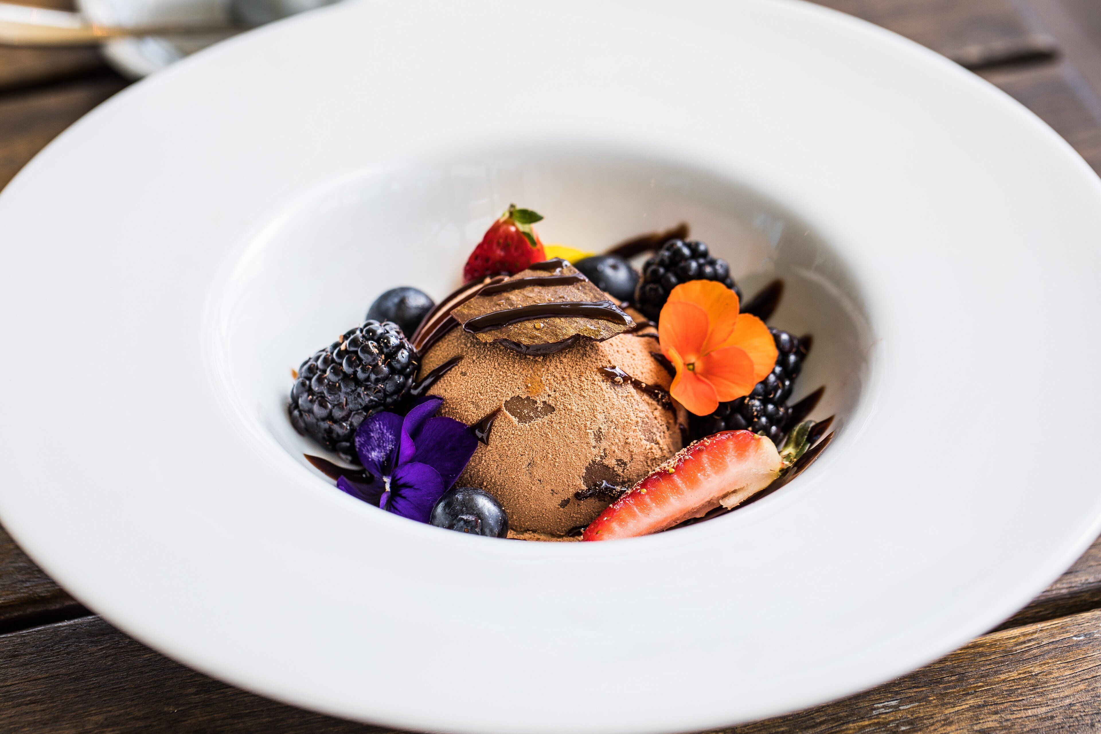 A dessert with chocolate ice cream, strawberries, blueberries, blackberries, and a flower on a white plate.