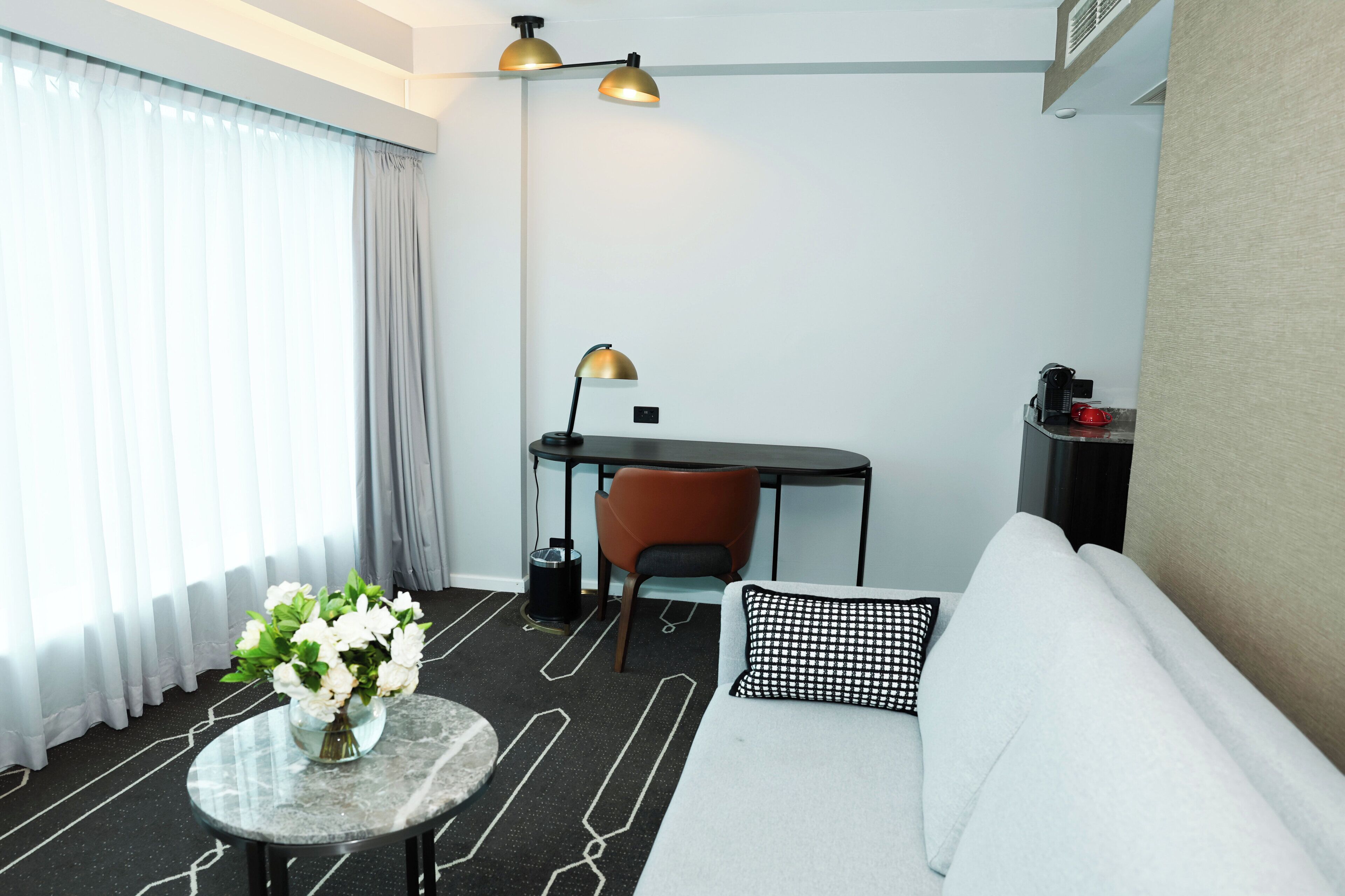 A modern hotel room with a sofa, a small round table with a vase of flowers, a desk with a chair, and two lamps.