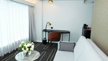 A modern hotel room with a sofa, a small round table with a vase of flowers, a desk with a chair, and two lamps.