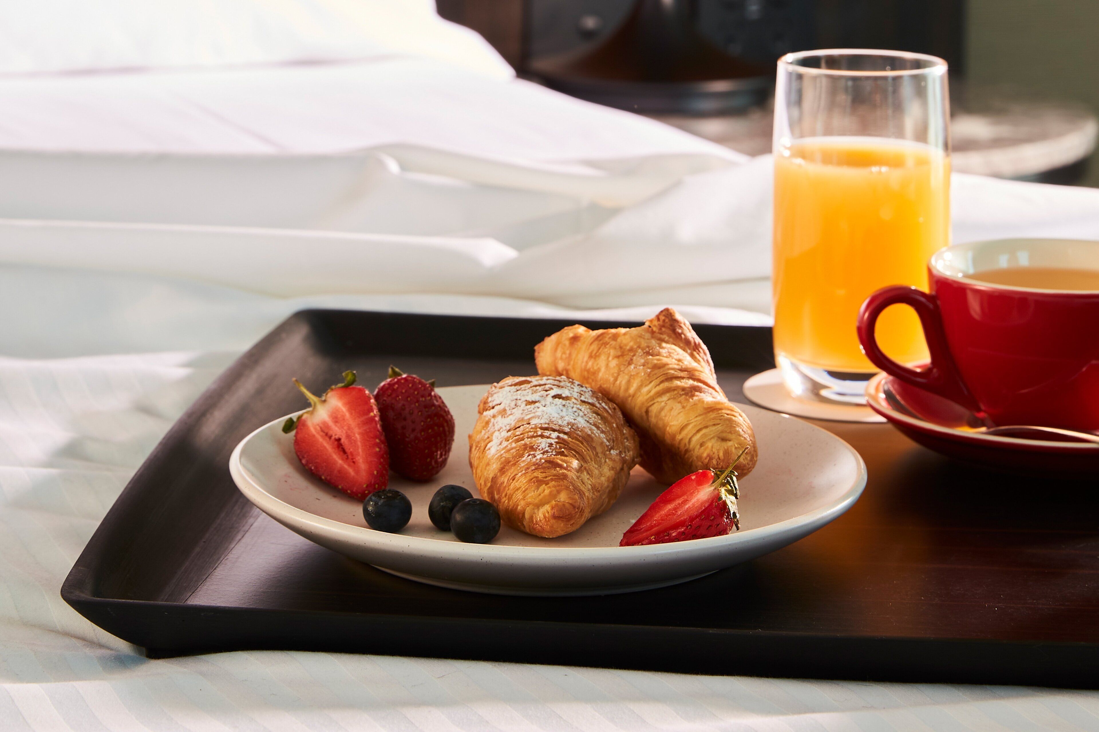 A breakfast tray with croissants, strawberries, blueberries, a glass of orange juice, and a red cup on a bed.