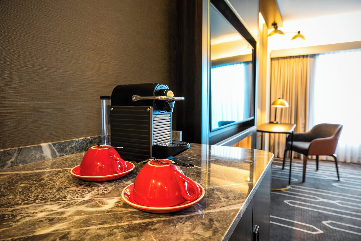 Ein Hotelzimmer mit Marmorplatte, einer Kaffeemaschine und zwei roten Tassen. Im Hintergrund stehen ein Sessel und ein Tisch.