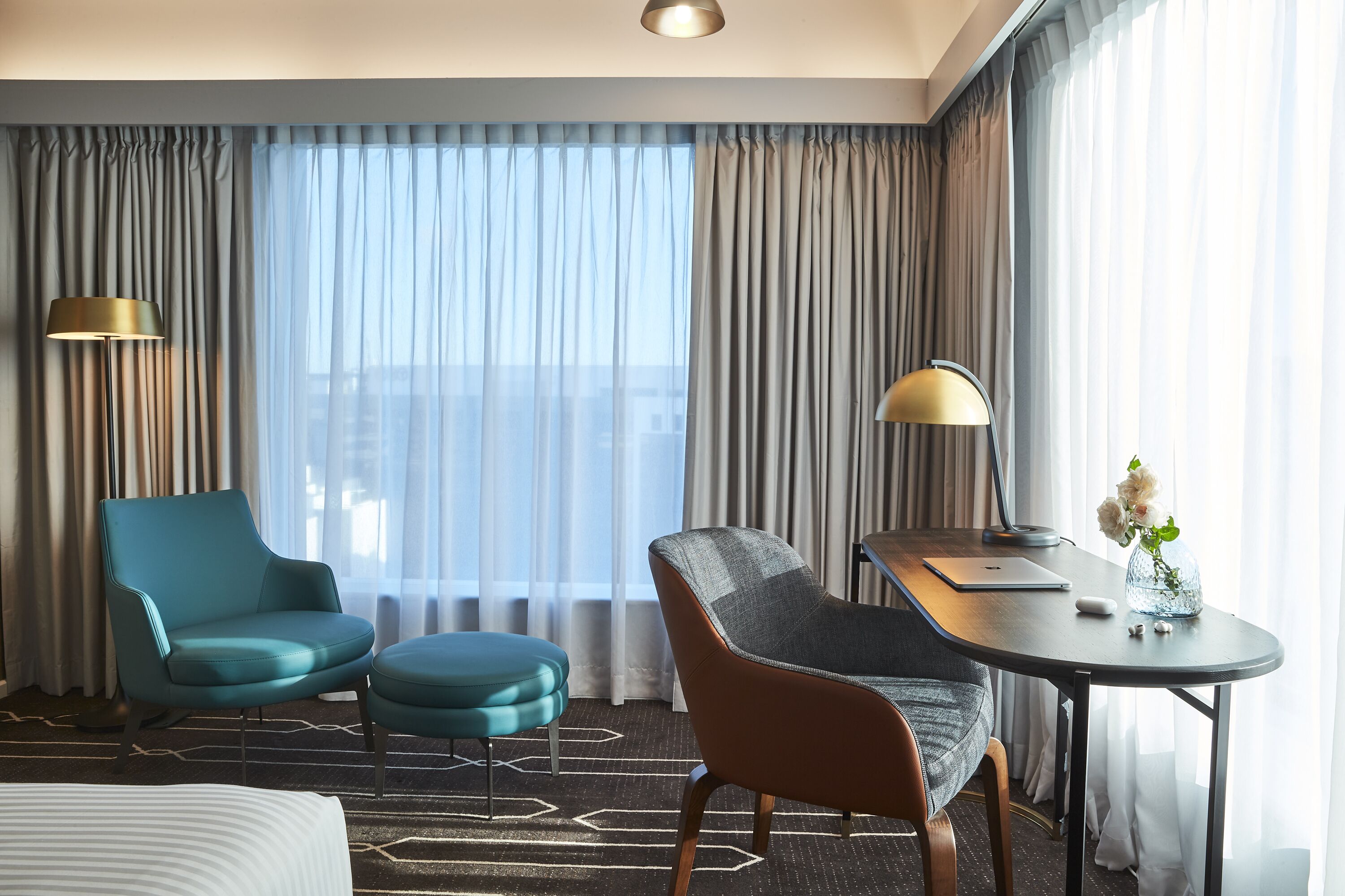 A hotel room with a bed, two chairs, a small table with a lamp, and a view of the city through large windows.