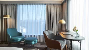 A hotel room with a bed, two chairs, a small table with a lamp, and a view of the city through large windows.