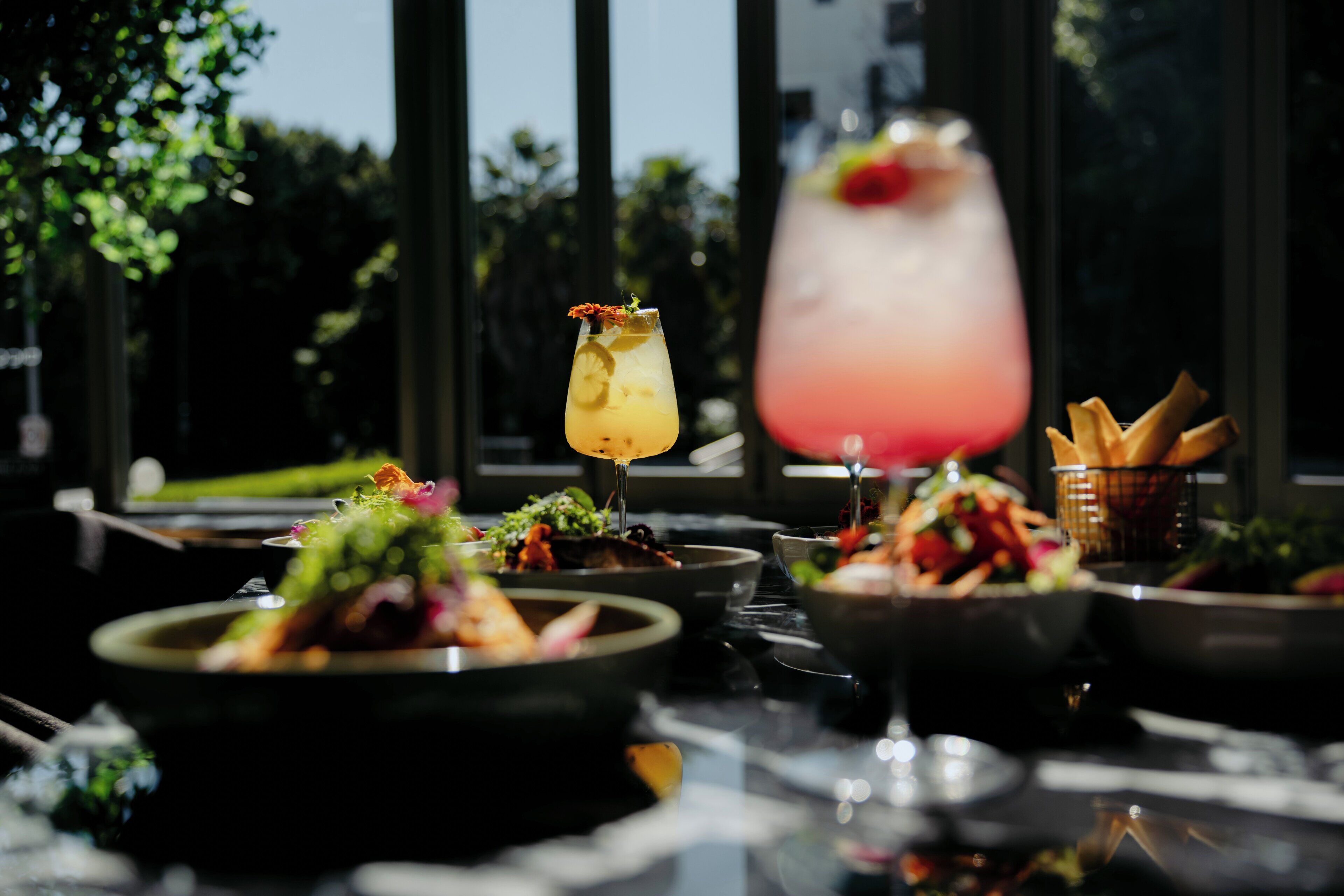 A table set with various dishes and drinks, with a view of trees and a building in the background.
