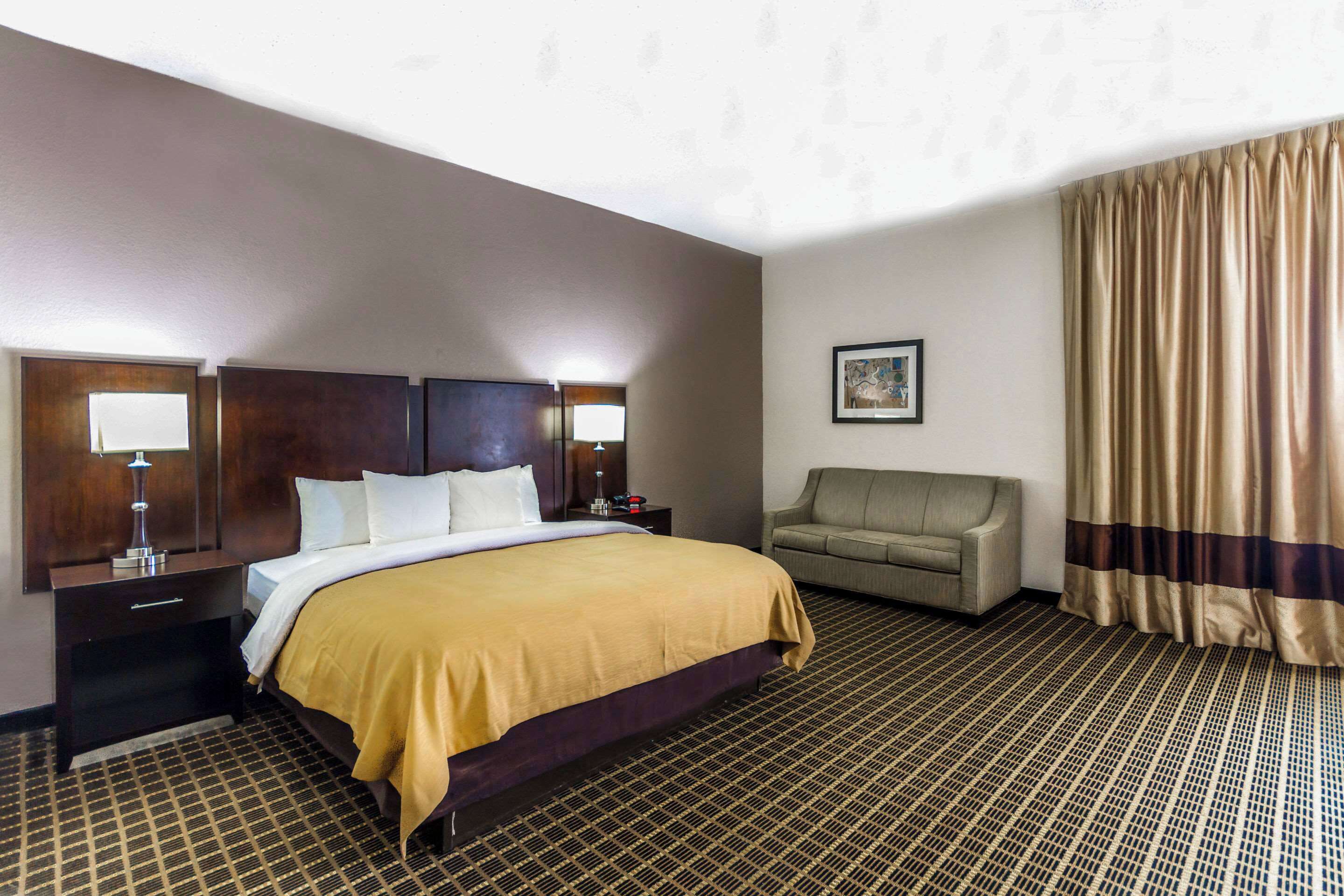 A hotel room with a bed, a nightstand, a lamp, a sofa, and a curtain.