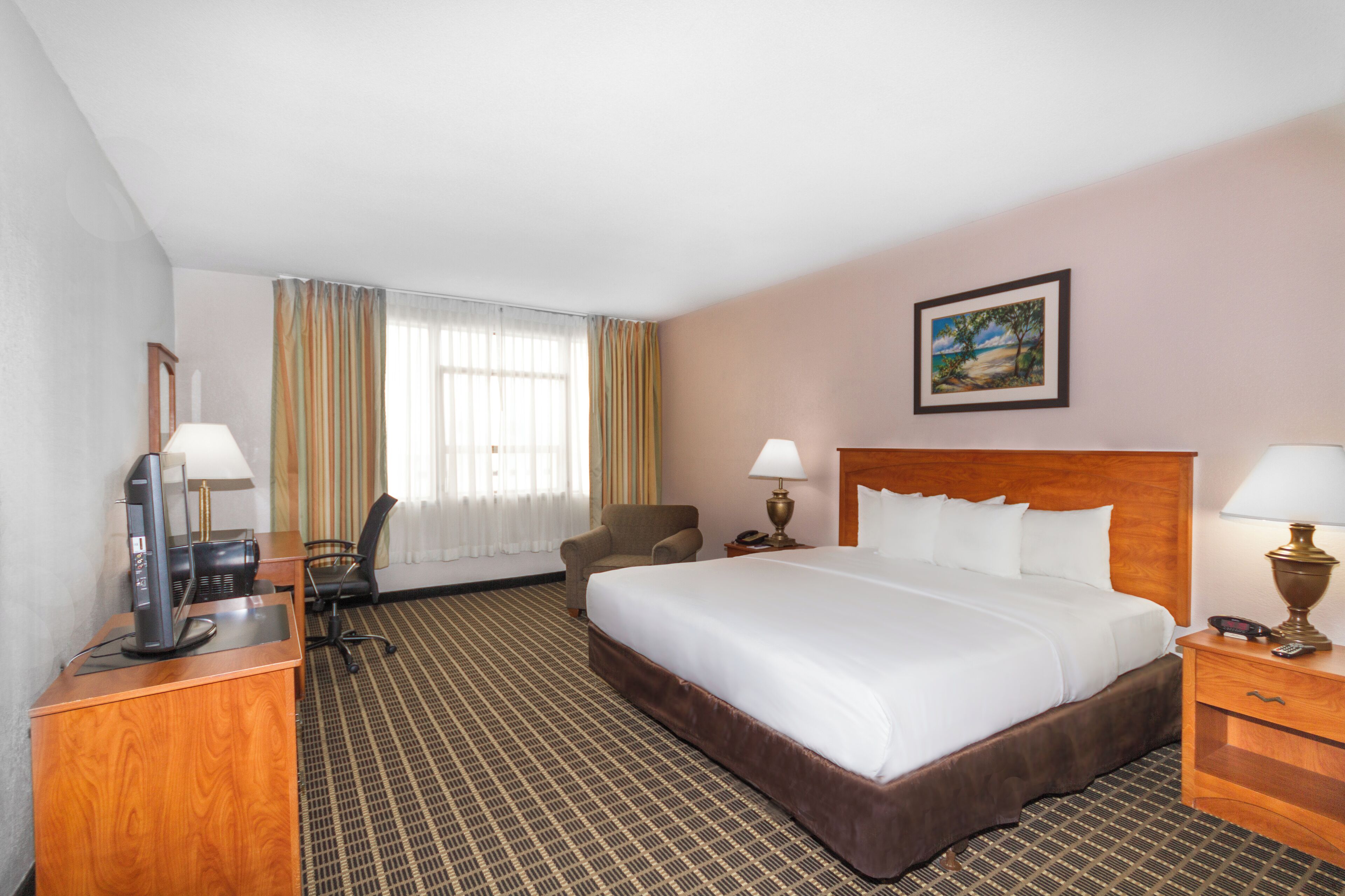 Ein Hotelzimmer mit einem großen Bett, einem Schreibtisch mit Computer, einem Stuhl und einem Nachttisch mit Lampe.