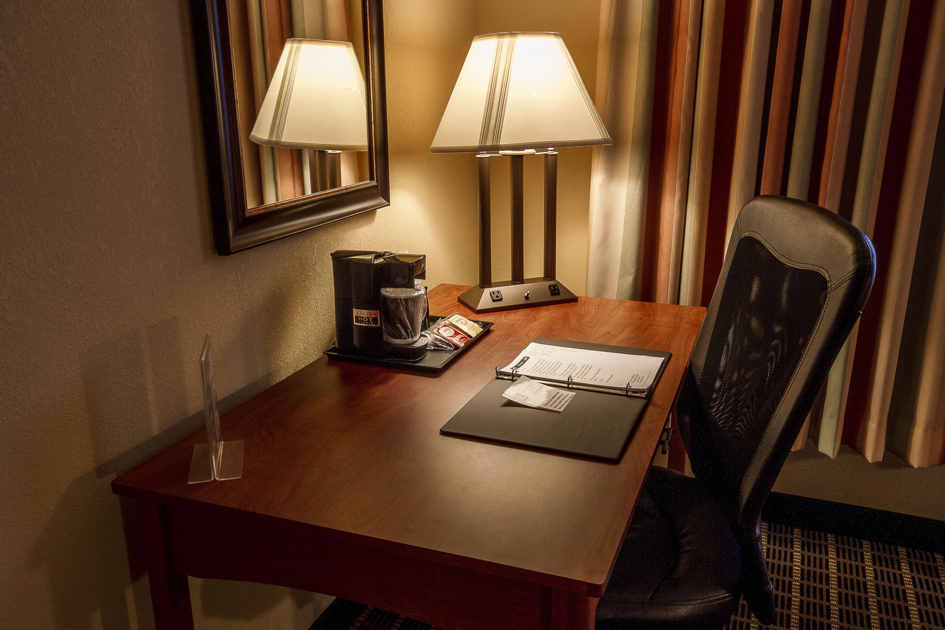A hotel room with a wooden desk, two lamps, a coffee maker, a black chair, and a mirror on the wall.