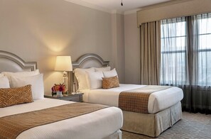 A hotel room with two beds, a nightstand with a lamp, a vase with flowers, and a large window with curtains.