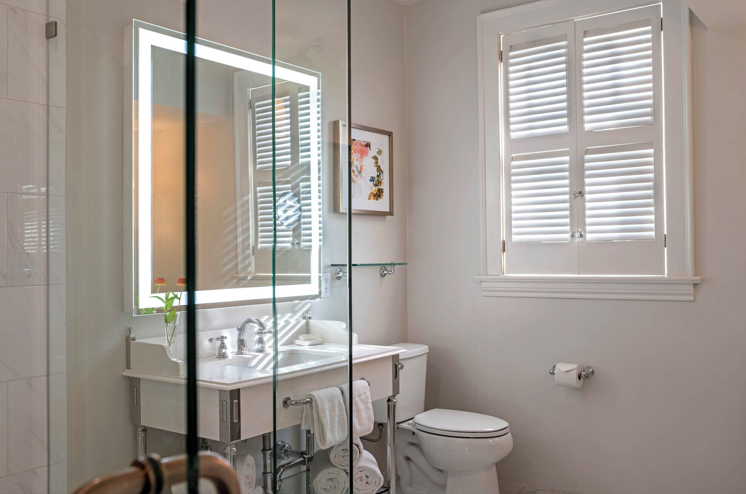 Un baño con cabina de ducha de vidrio, un lavamanos blanco, inodoro y ventana con contraventanas.