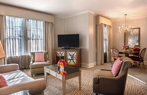 A hotel room with a TV, two sofas, a dining table with chairs, and a chandelier.