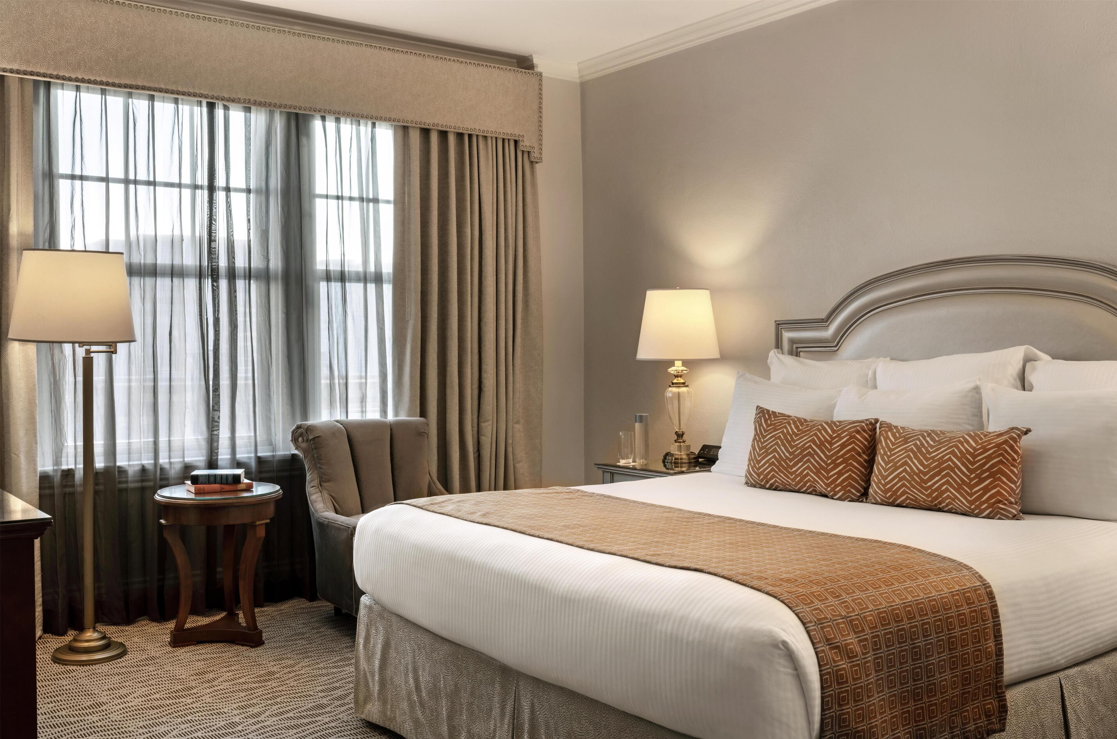 A hotel room with a large bed, two bedside lamps, a chair, a small table with books, and a window with curtains.