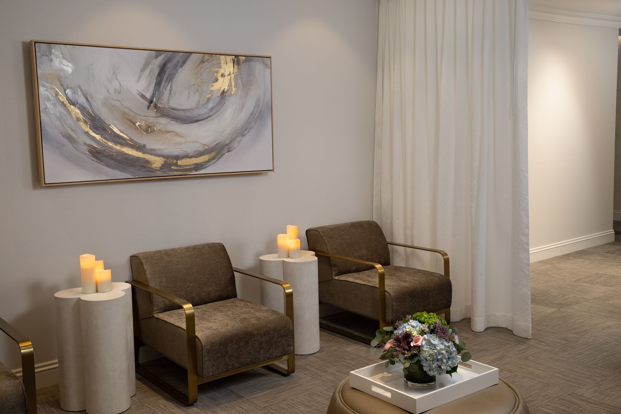 A modern room with two armchairs, a white side table with candles, a large abstract painting, and a floral arrangement on a tray.