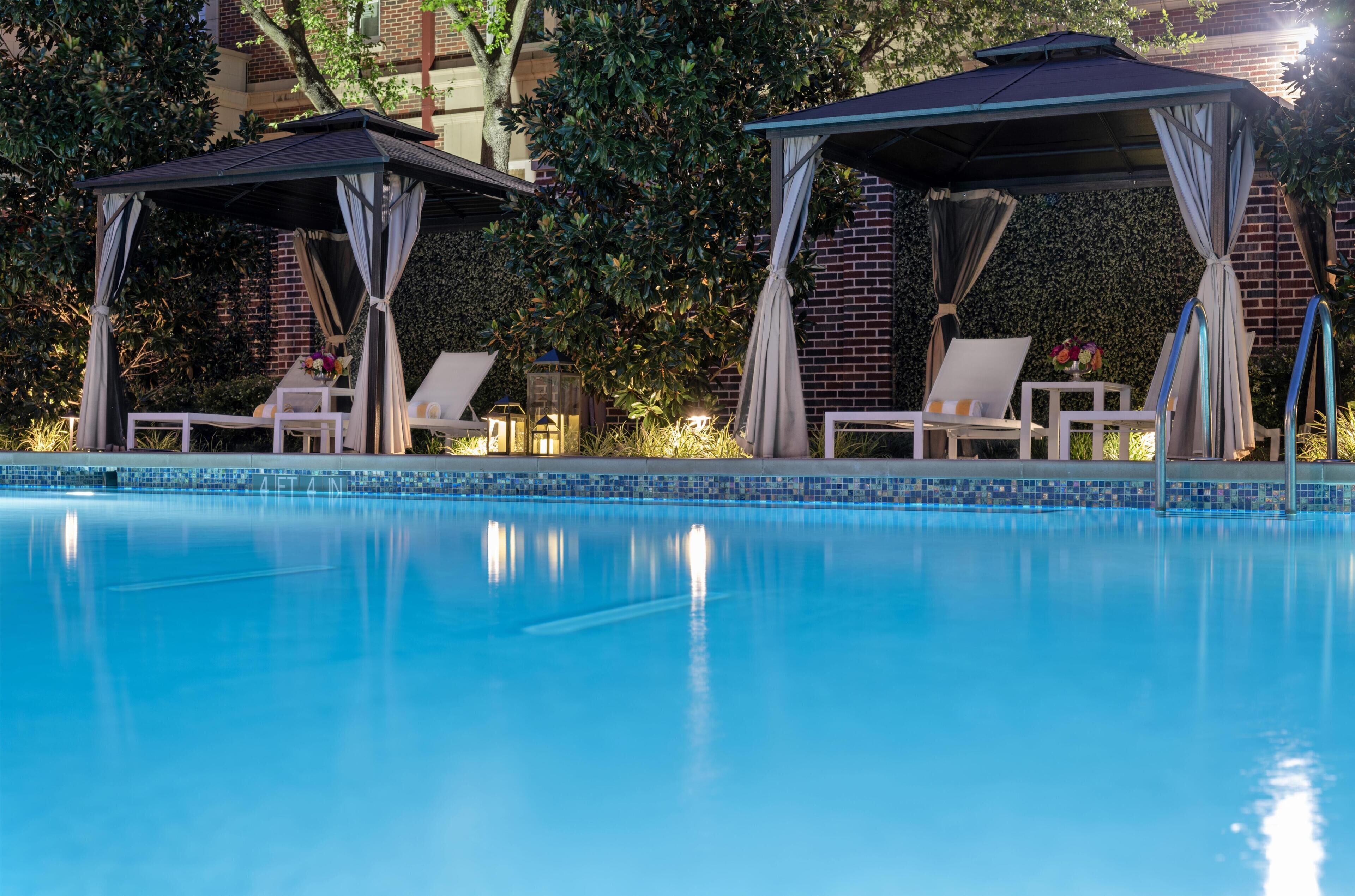 Una piscina con sillones de descanso y gazebos, rodeada de vegetación y un muro de ladrillo.