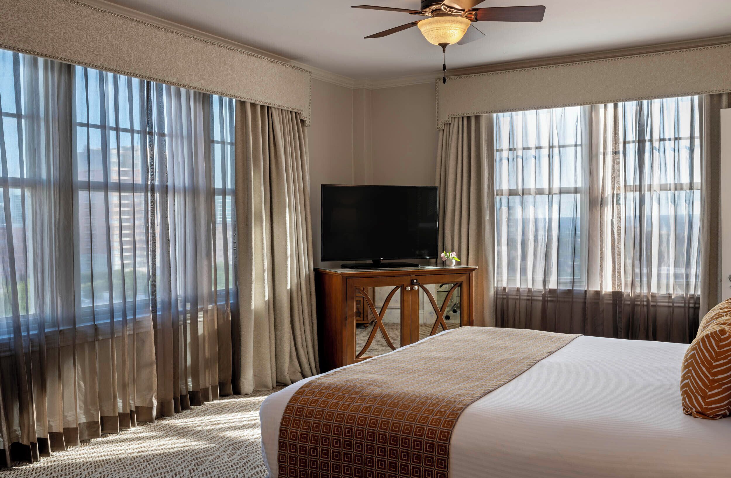 A hotel room with a bed, a television on a wooden table, and large windows with curtains.