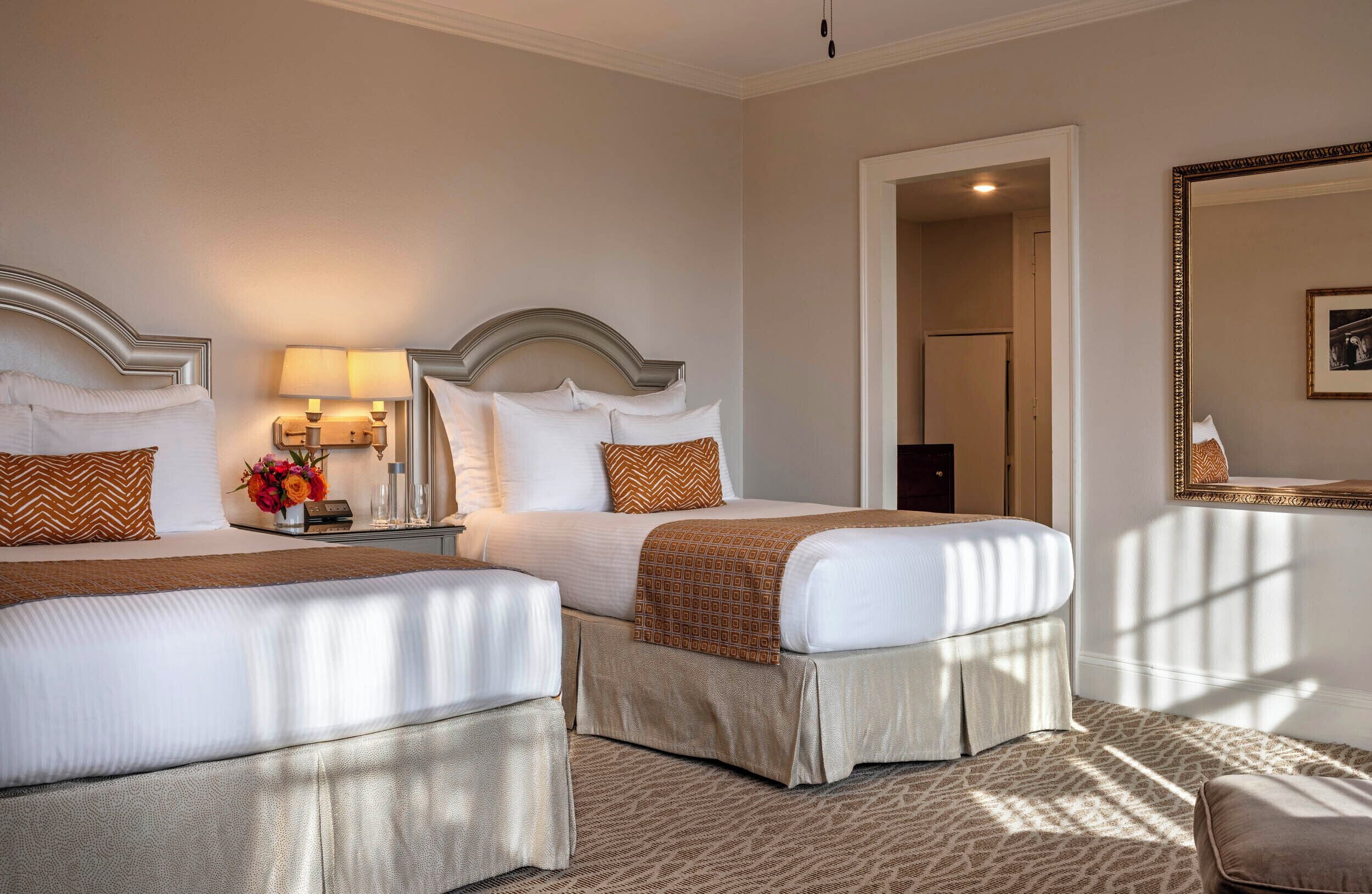 A hotel room with two beds, a nightstand with a lamp, a mirror, and a framed picture on the wall.
