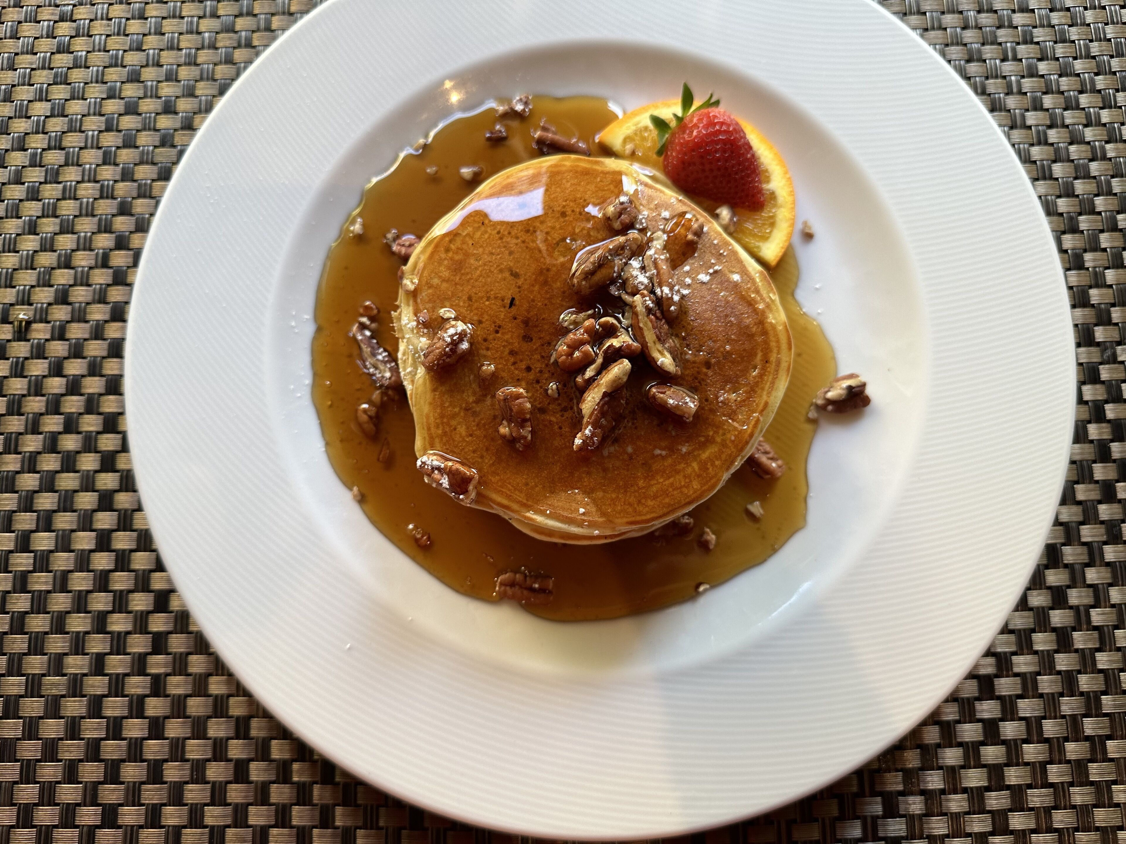 A plate with pancakes topped with pecans and syrup, accompanied by a strawberry and orange slice.