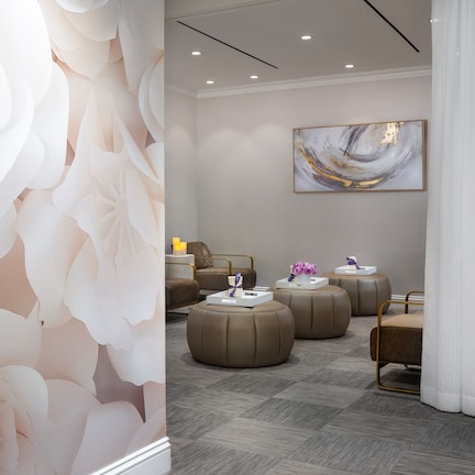 A modern hotel room with a large floral wall mural, a comfortable seating area with ottomans, a large abstract painting on the wall, and a neatly arranged side table with a lamp.