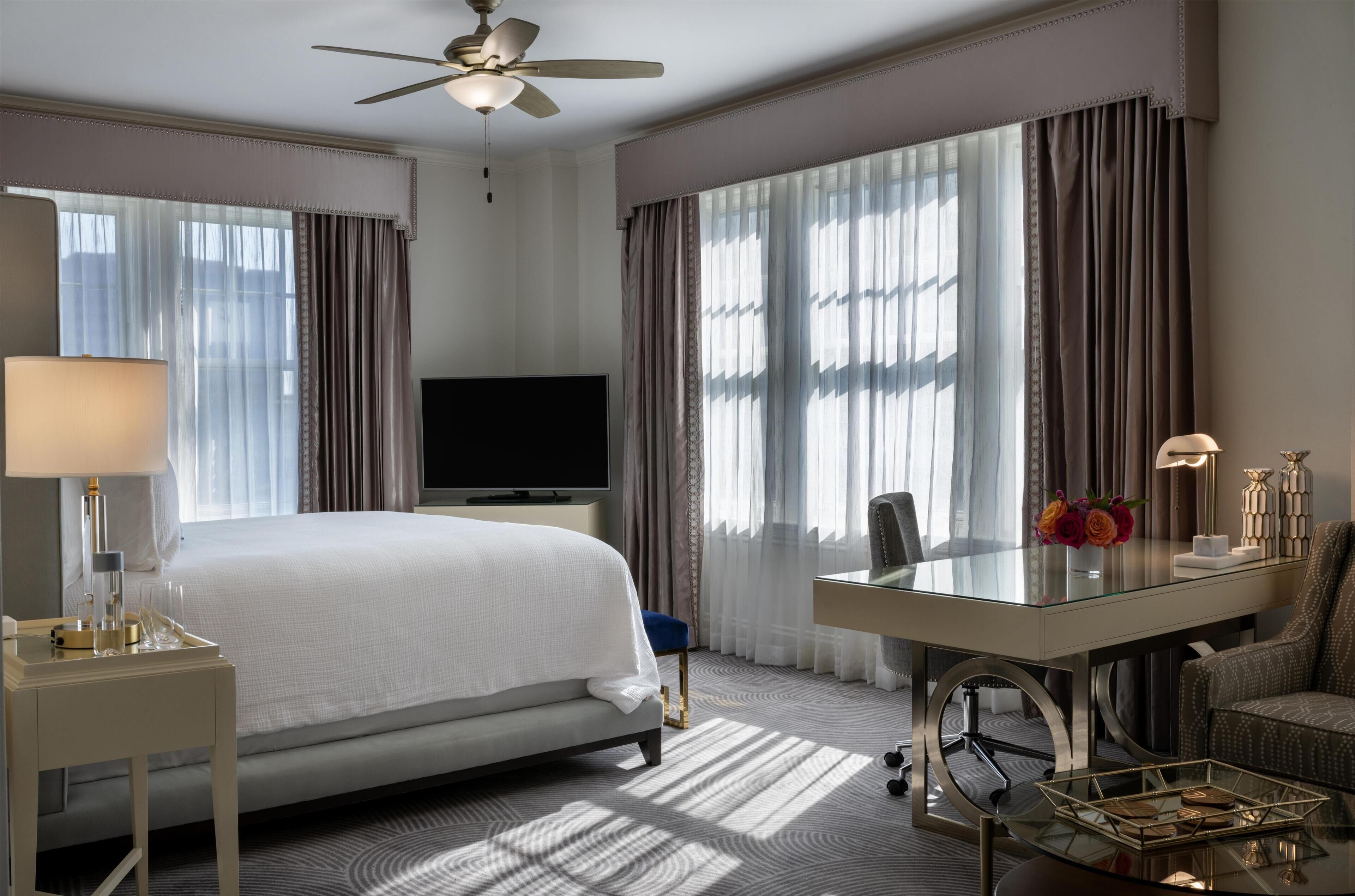 A hotel room with a bed, a desk, a chair, a television, and a ceiling fan.