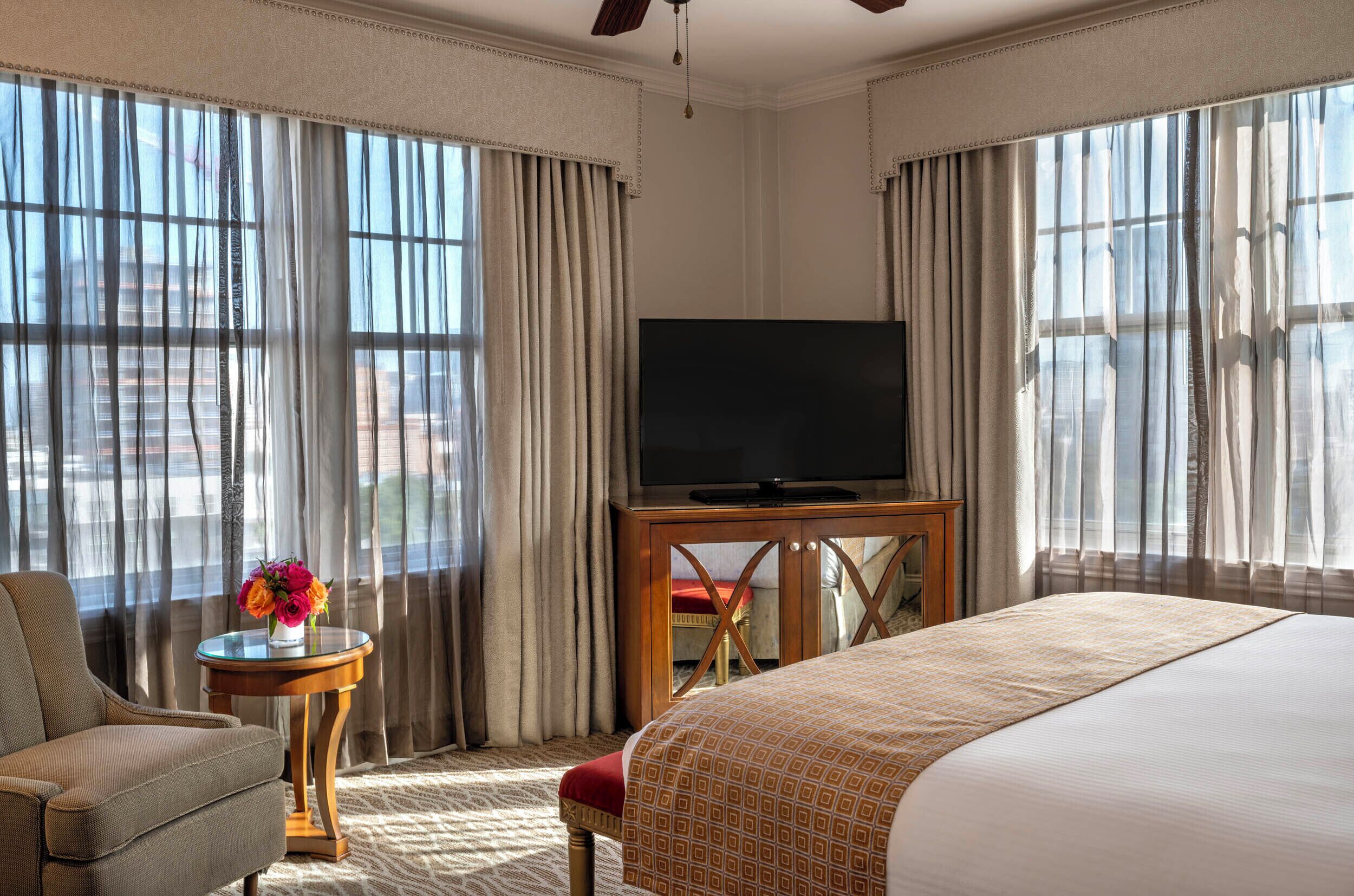 A hotel room with a bed, a TV on a wooden stand, a chair, a small table with a vase of flowers, and large windows with curtains.