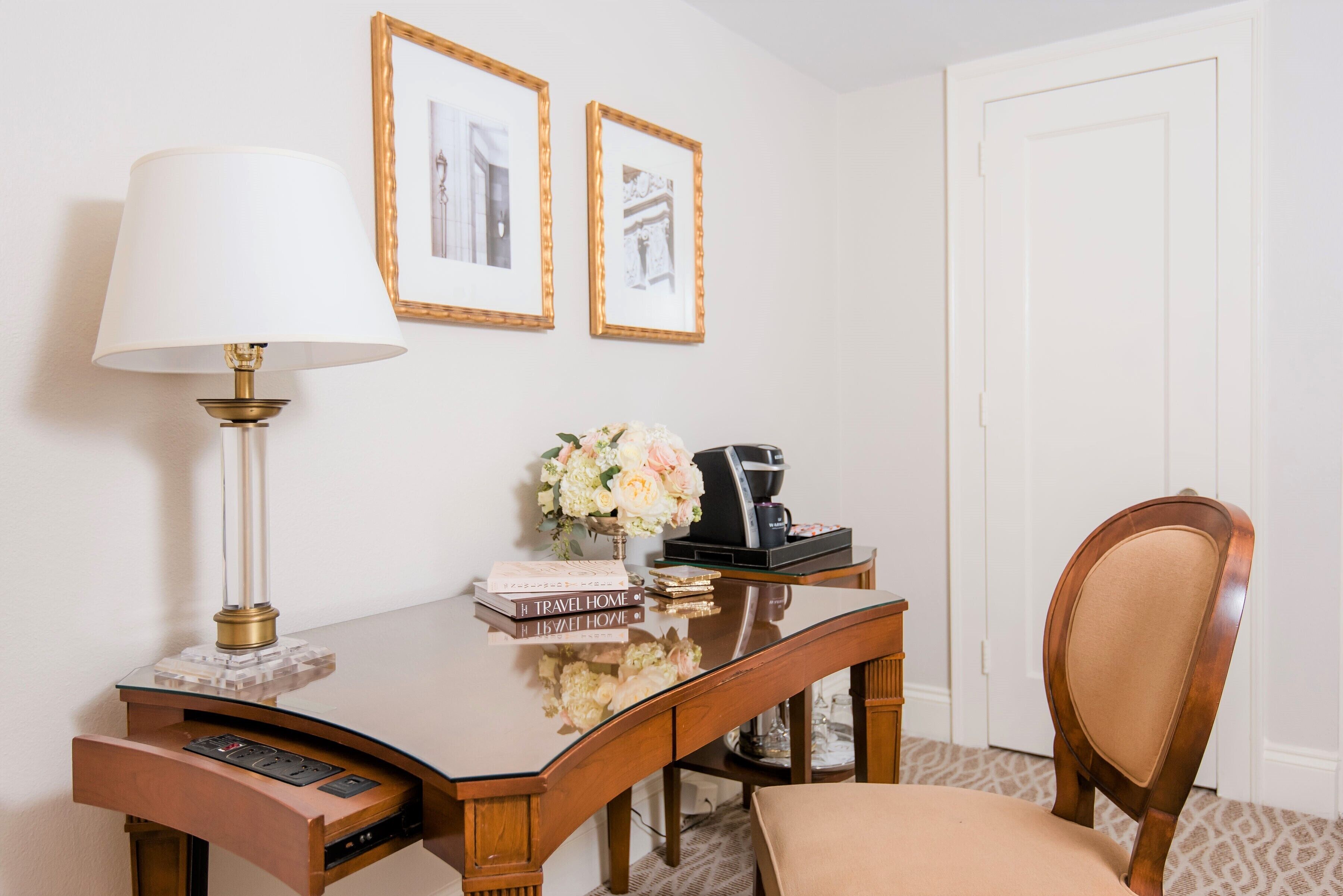 Habitación de hotel con un escritorio de madera, una lámpara, cuadros en la pared, un florero con flores y una silla.