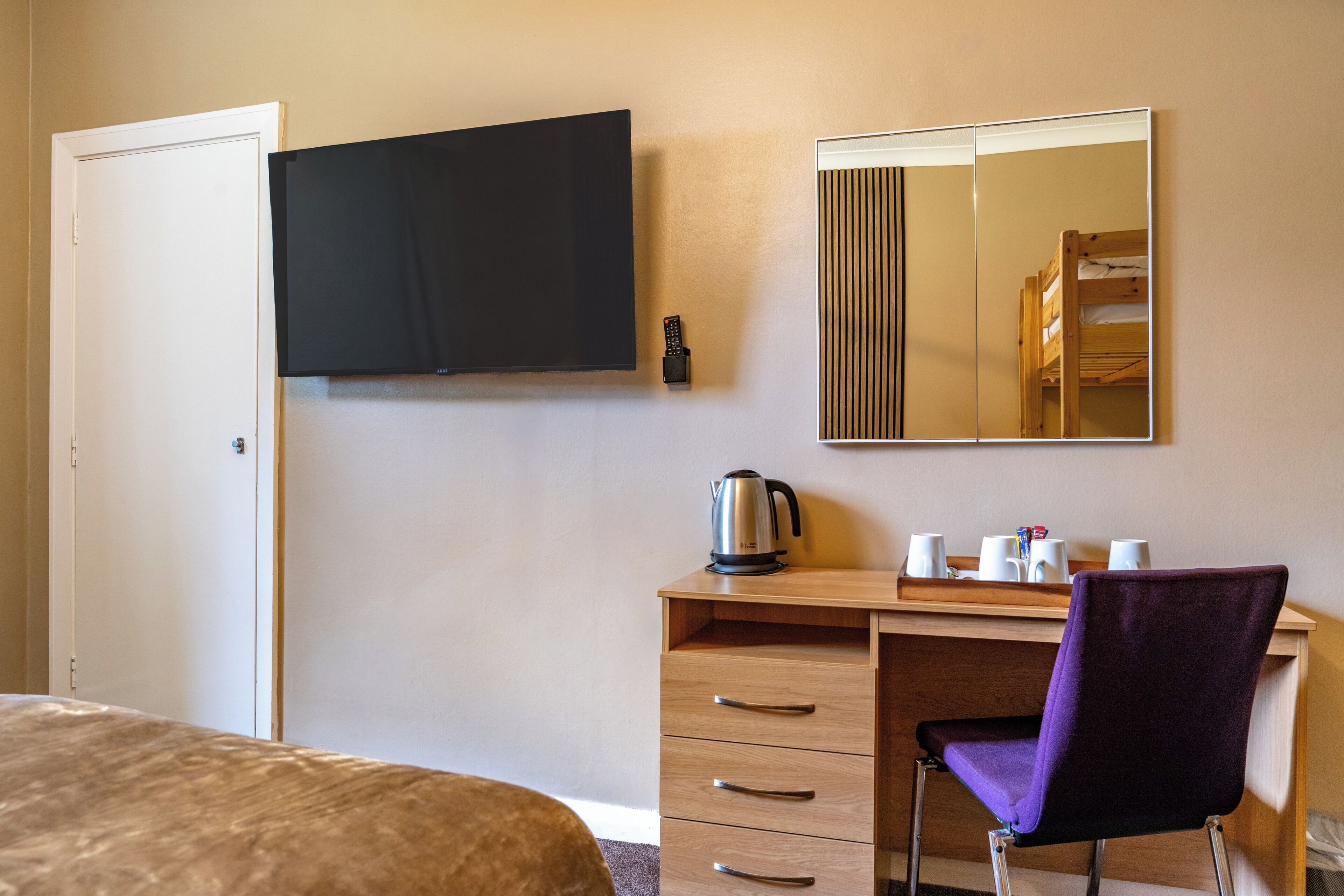 A hotel room with a bed, a desk with a chair, a television, a mirror, and a kettle.