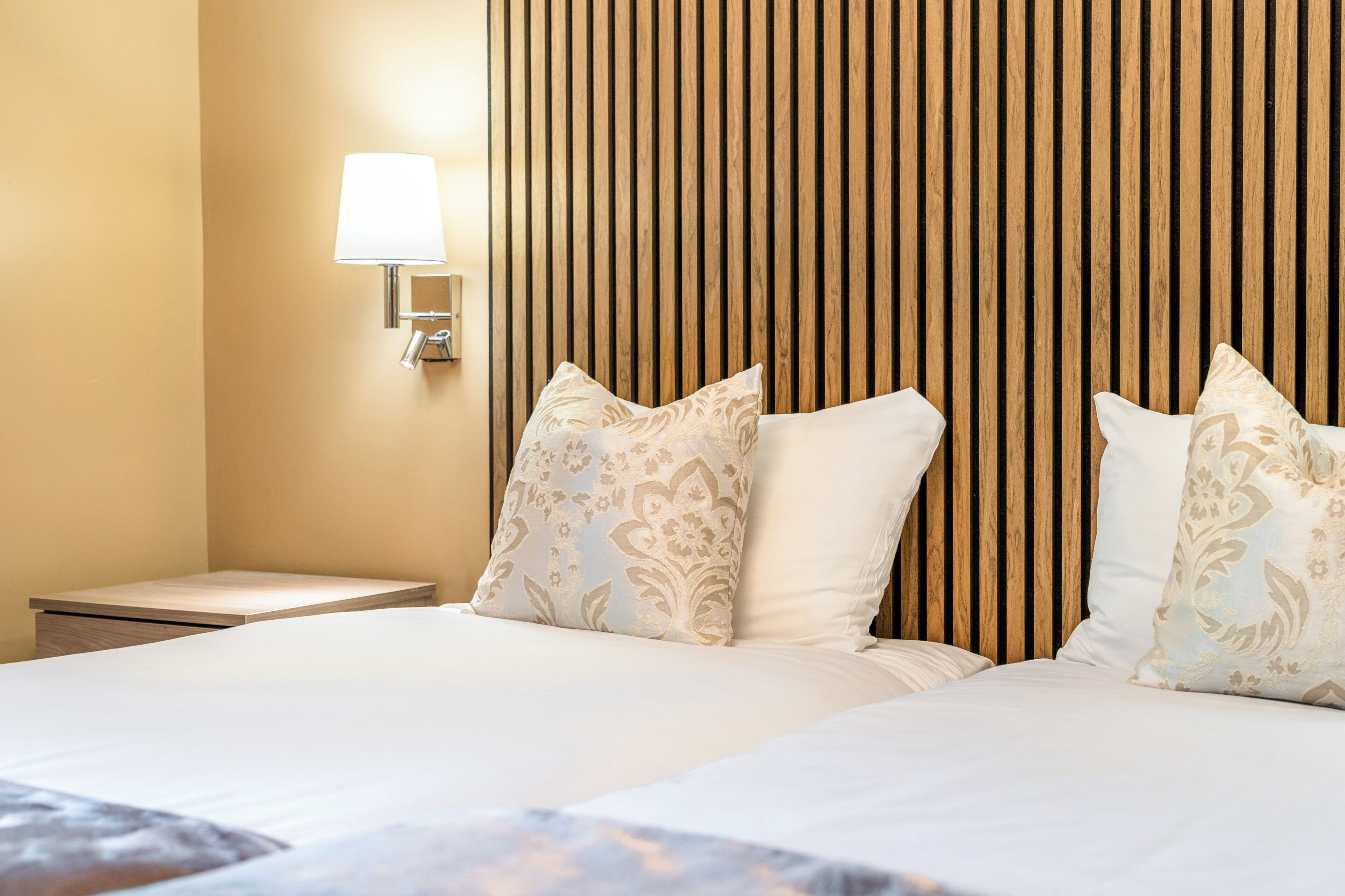 A hotel room with a bed, two pillows, a nightstand, and a wall-mounted lamp.