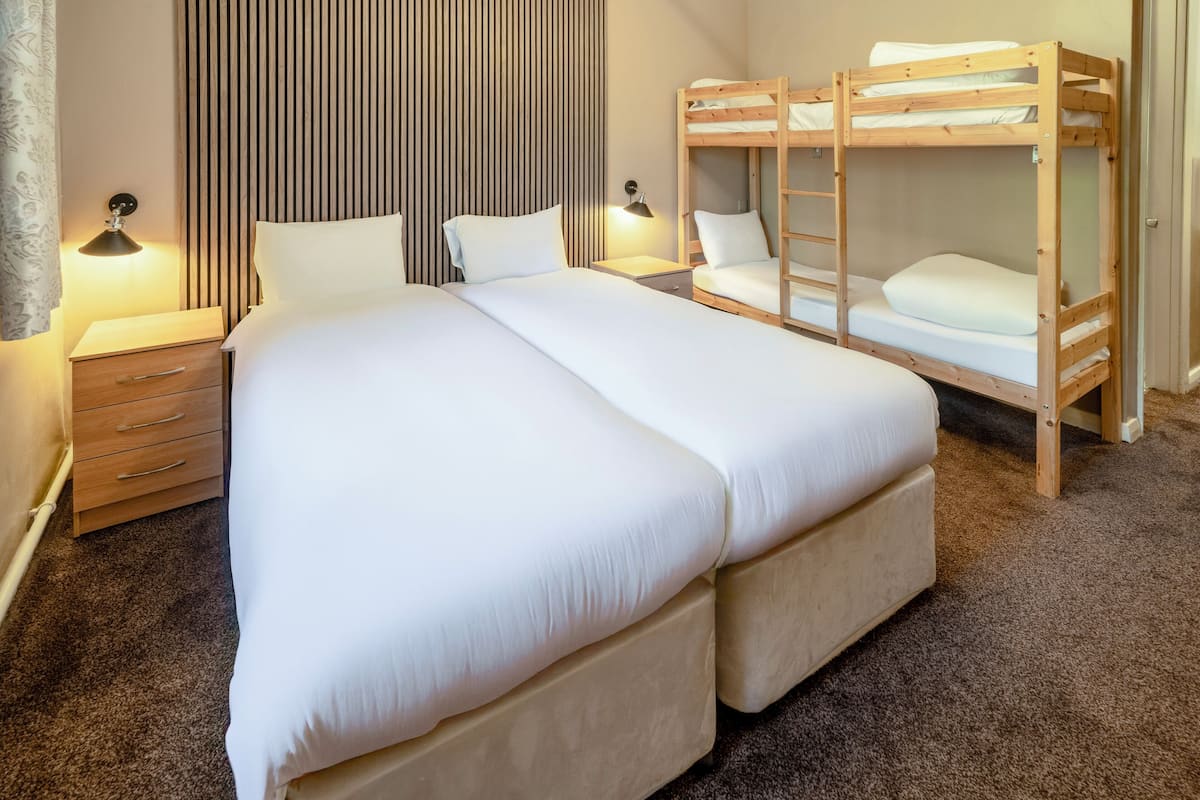 A double bed with a wooden headboard, a nightstand with a lamp, a dresser, and a bunk bed with a ladder.