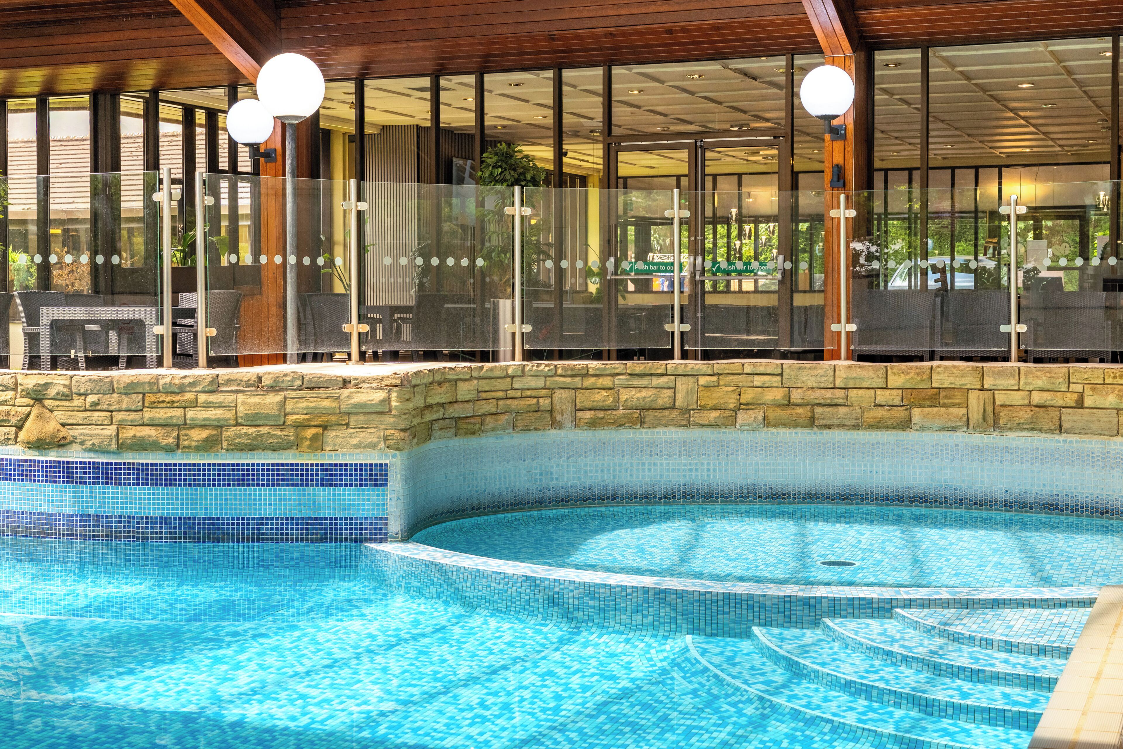 Una piscina interior con agua cristalina y azul, rodeada por un muro de piedra y paneles de vidrio.