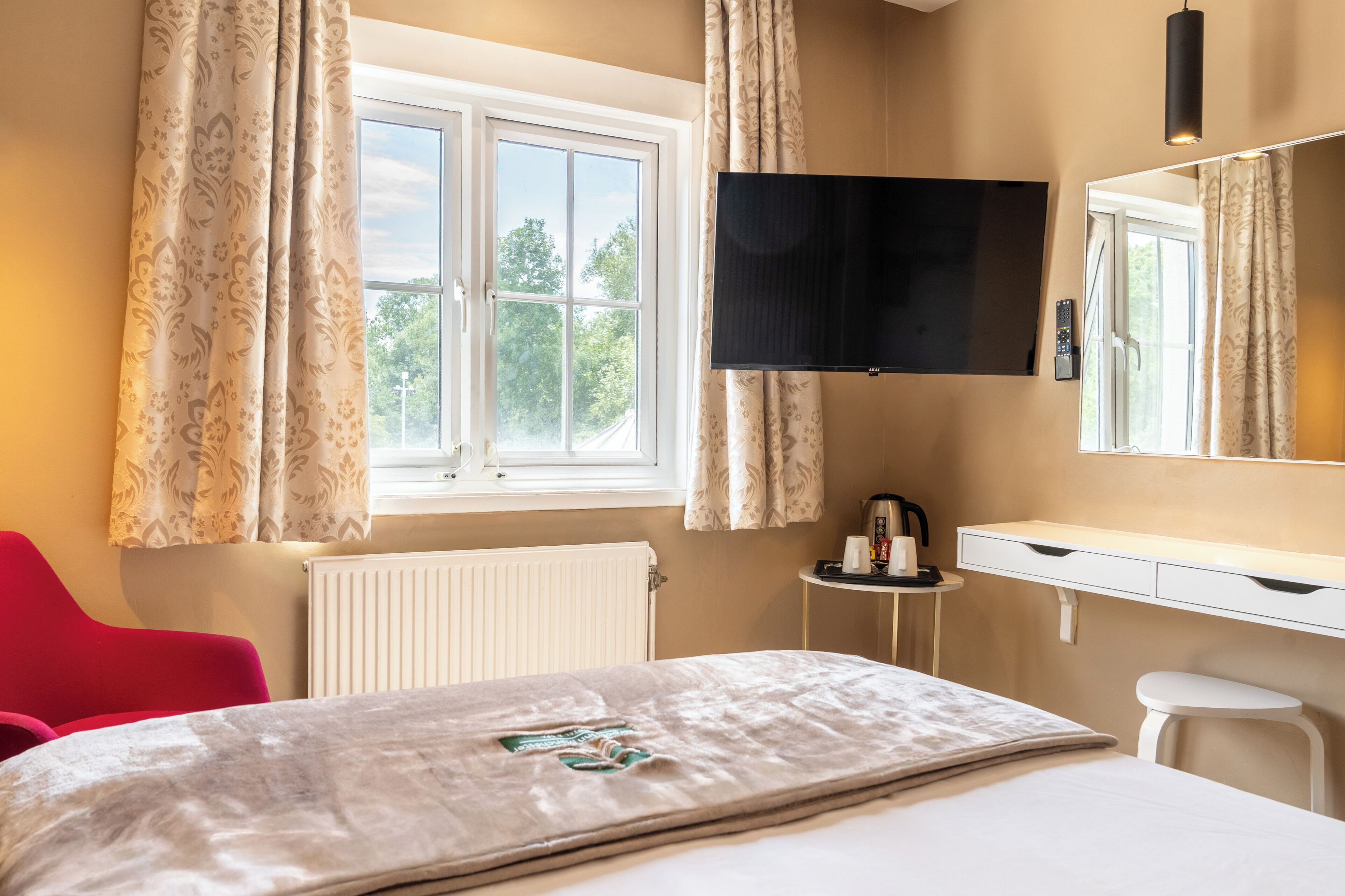 A hotel room with a bed, a desk, a chair, a radiator, a window with curtains, and a flat-screen TV mounted on the wall.