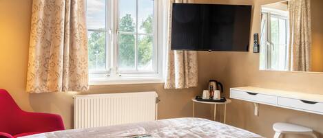 A hotel room with a bed, a desk, a chair, a radiator, a window with curtains, and a flat-screen TV mounted on the wall.