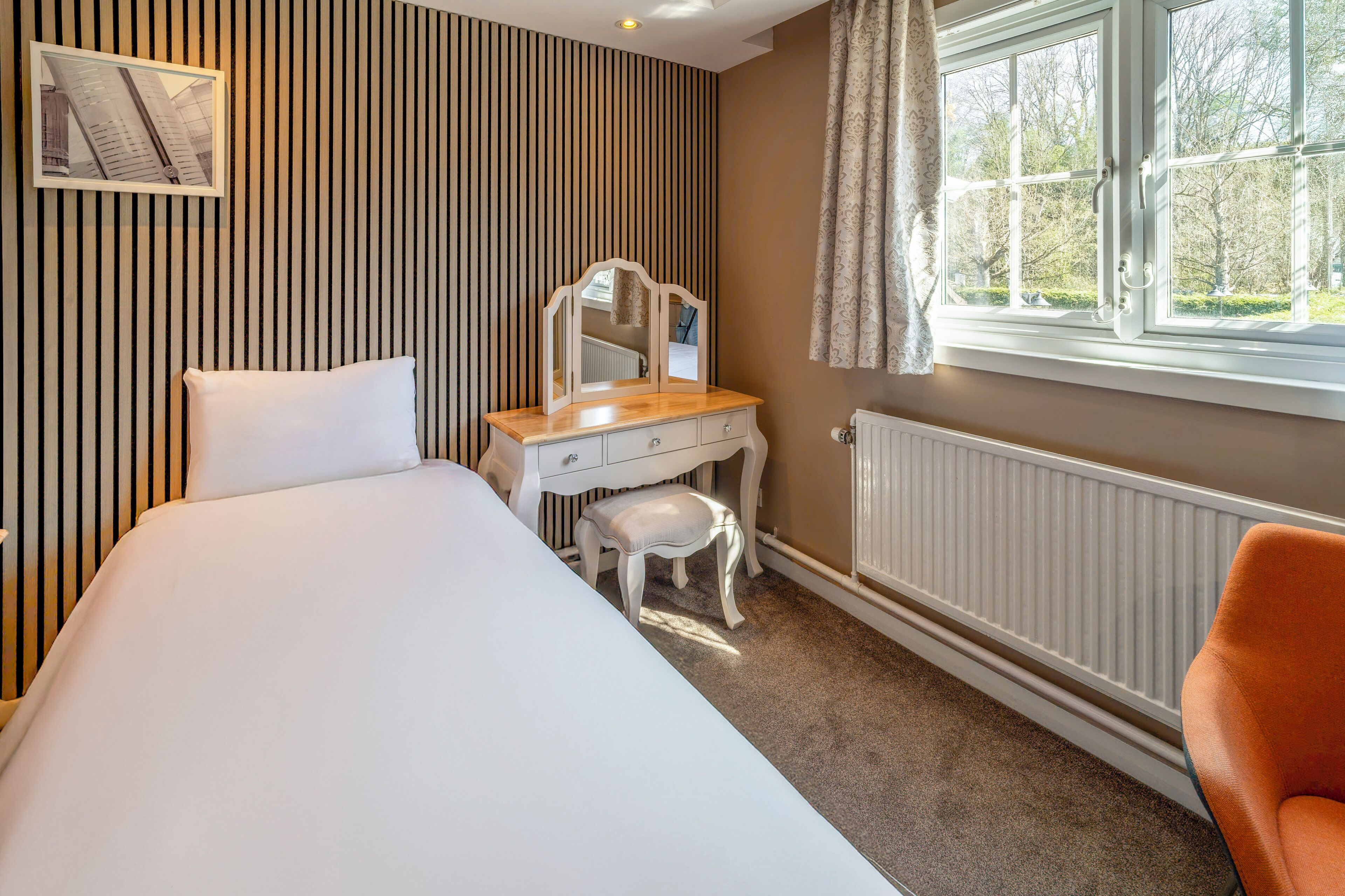 A bedroom with a bed, a vanity with a mirror, a chair, and a window with a view of trees.