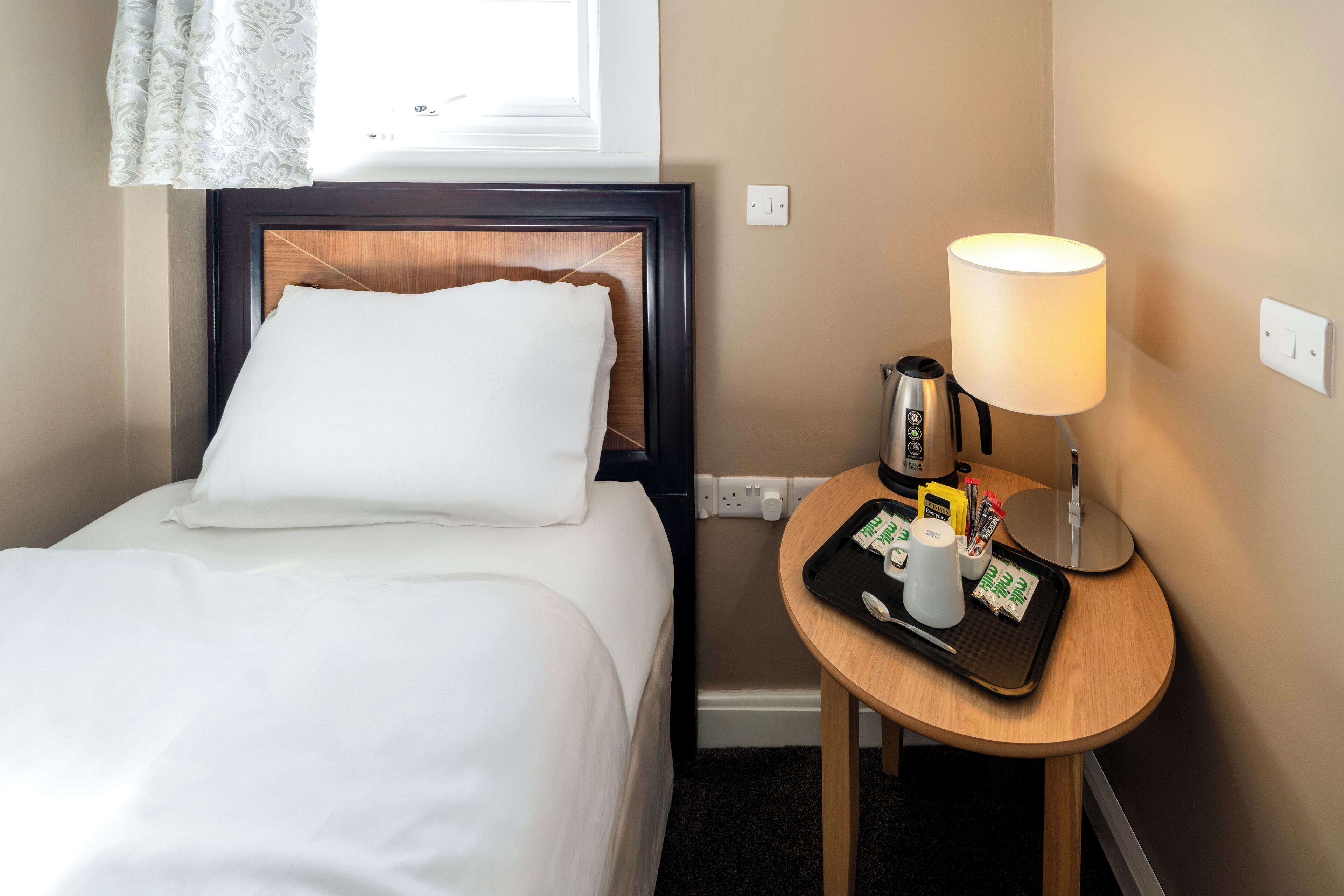 A hotel room with a neatly made bed, a bedside table with a lamp, a tray with toiletries, and a window with a curtain.