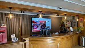 A hotel reception area with a wooden reception desk, a mounted television displaying news, a red telephone, and a plant. - Britannia Airport Inn Manchester (Wilmslow)