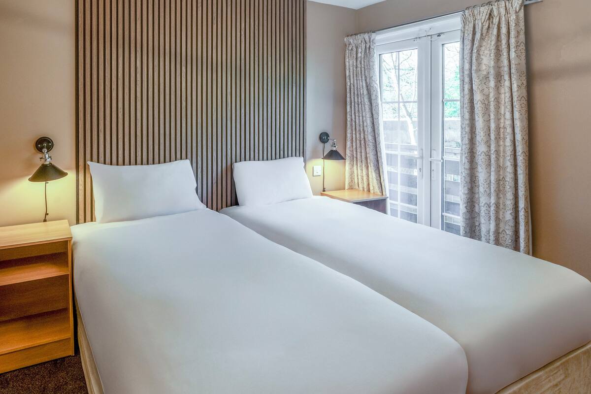 A hotel room with two single beds, a wooden nightstand, a lamp, and a window with curtains.