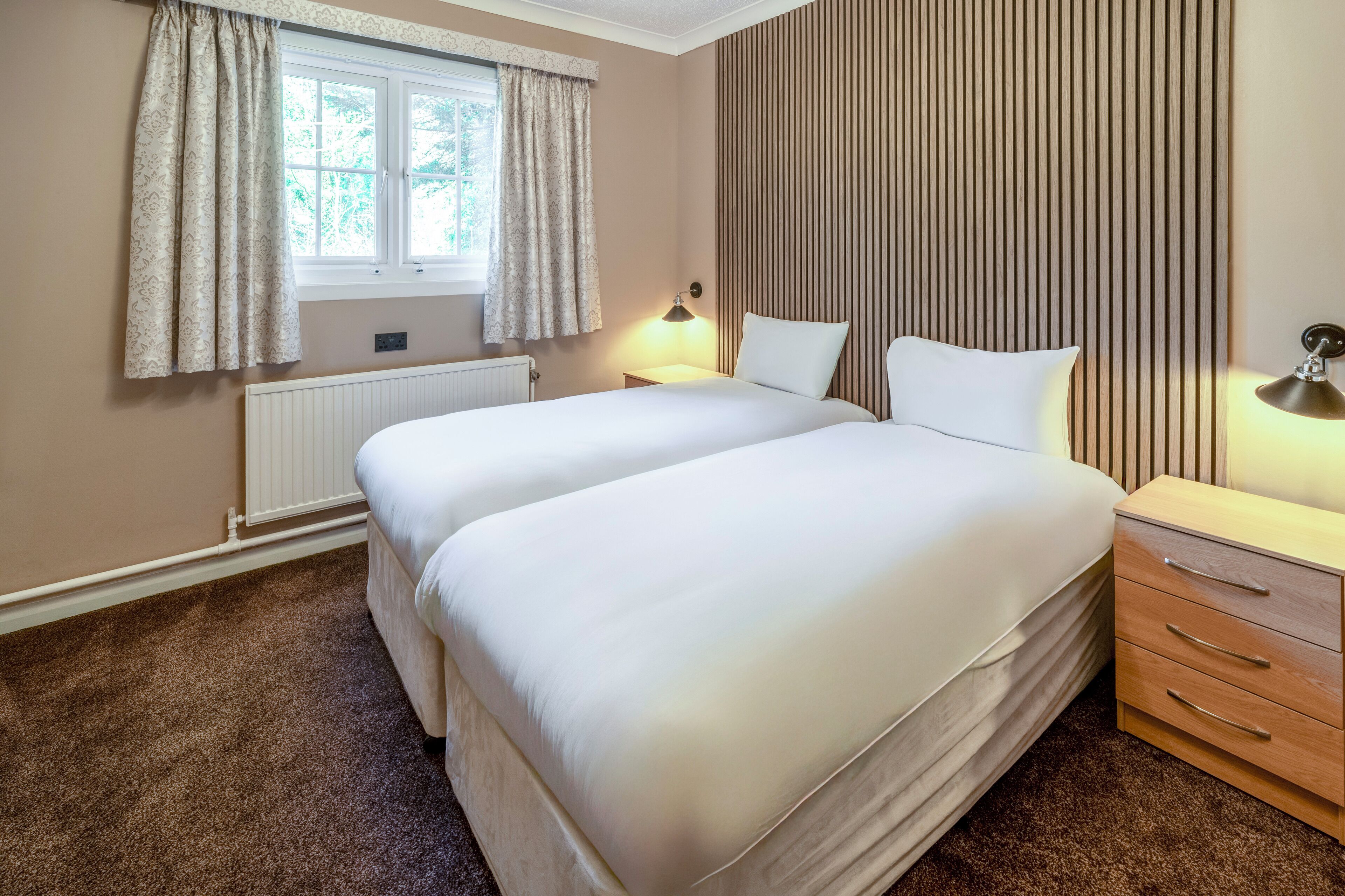 A hotel room with two beds, a window with curtains, a radiator, a bedside lamp, and a wooden nightstand with drawers.